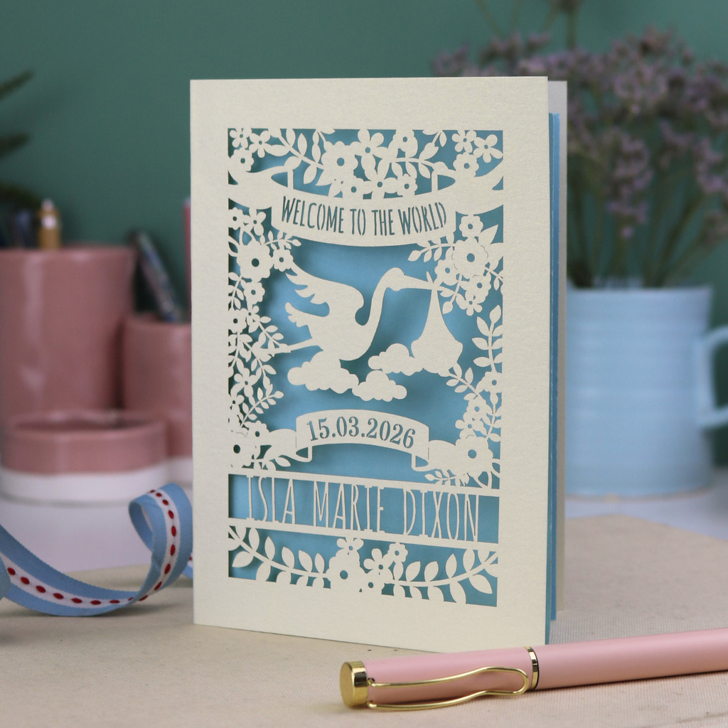 A personalised new baby card with laser cut floral stork design and "Welcome to the World" in cut out text in a banner, with the baby's date of birth and name