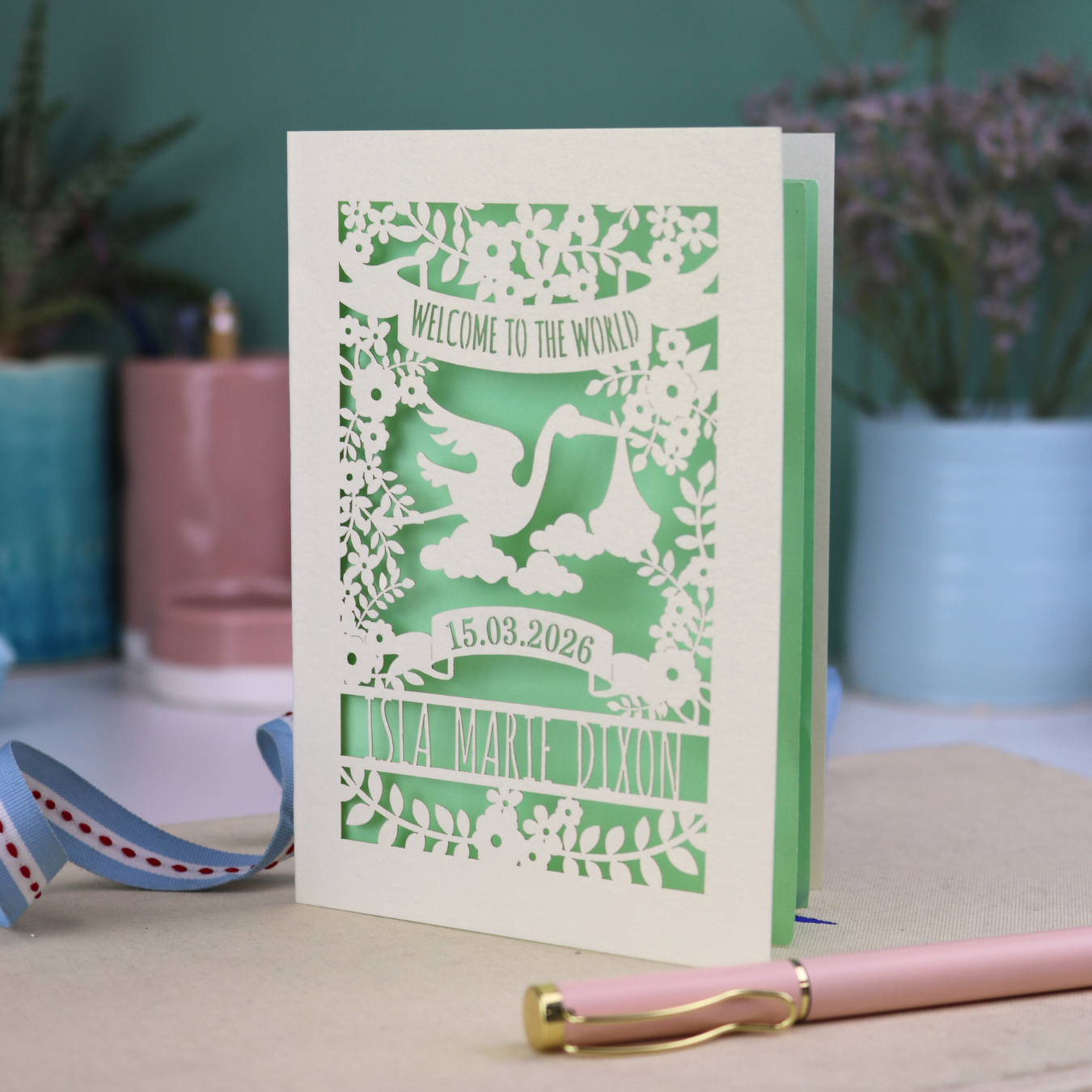 A personalised new baby card with laser cut floral stork design and "Welcome to the World" in cut out text in a banner, with the baby's date of birth and name