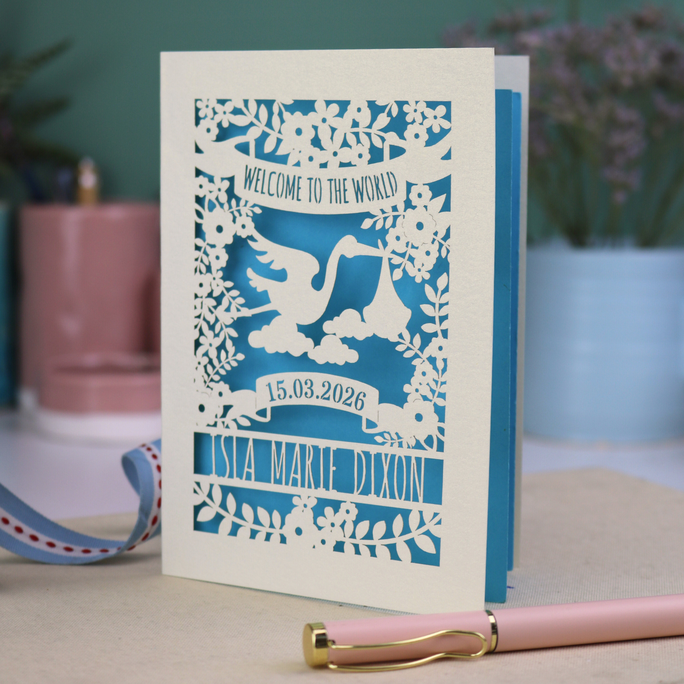 A personalised new baby card with laser cut floral stork design and "Welcome to the World" in cut out text in a banner, with the baby's date of birth and name