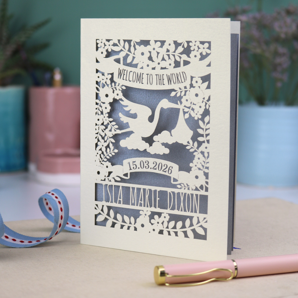 A personalised new baby card with laser cut floral stork design and "Welcome to the World" in cut out text in a banner, with the baby's date of birth and name