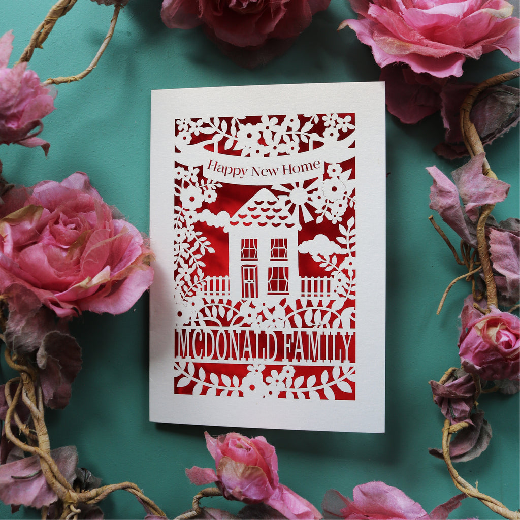 A personalised new home card, laser cut and featuring a house surrounded by flowers. There is a banner with the words "Happy new home" and a line of text at the bottom of the card