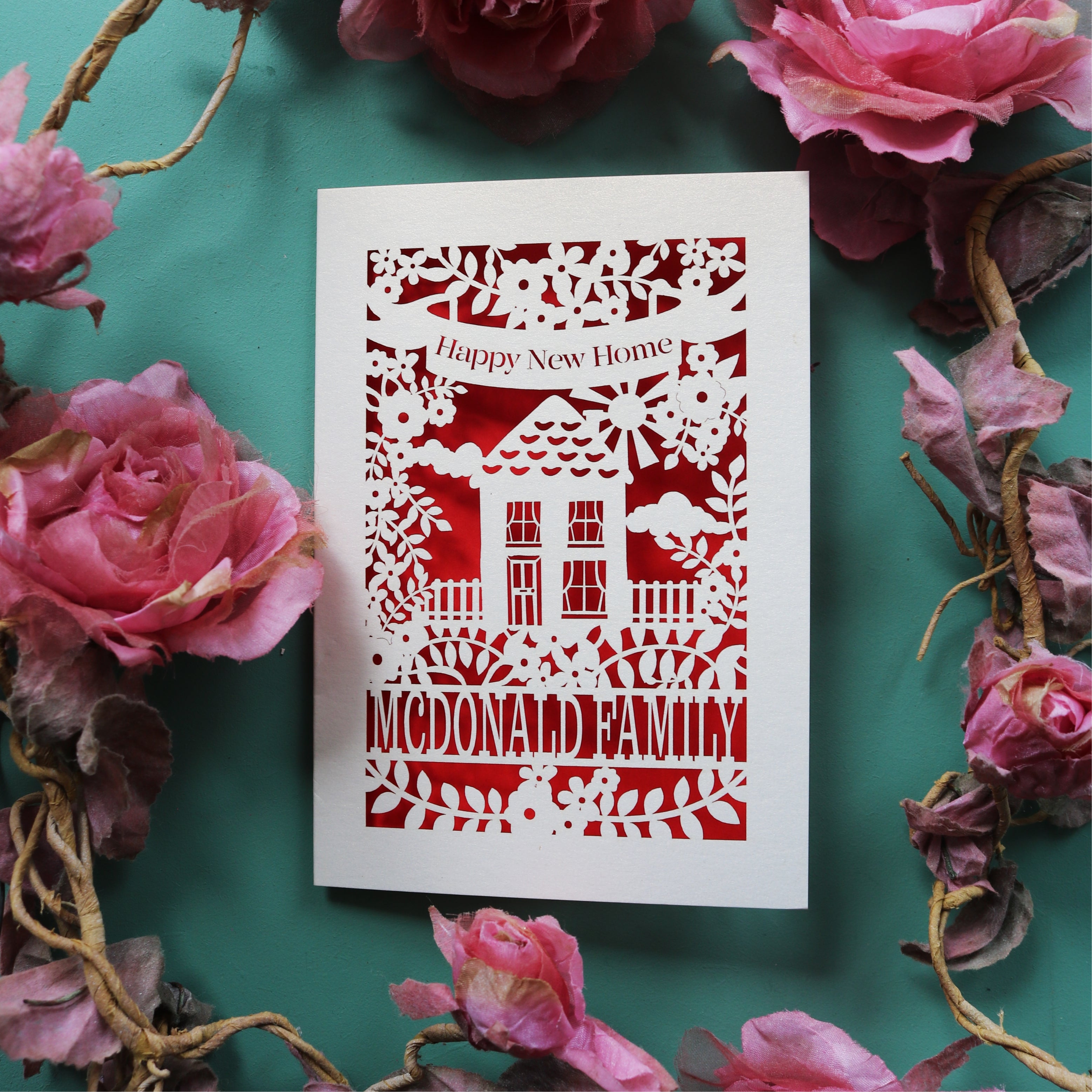 A personalised new home card, laser cut and featuring a house surrounded by flowers. There is a banner with the words "Happy new home" and a line of text at the bottom of the card