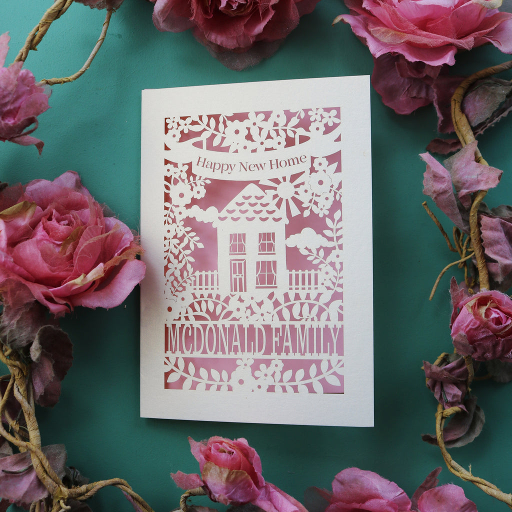 A cream and light pink paper cut house warming card with a house surrounded by flowers. This card reads "happy new home" in a banner 