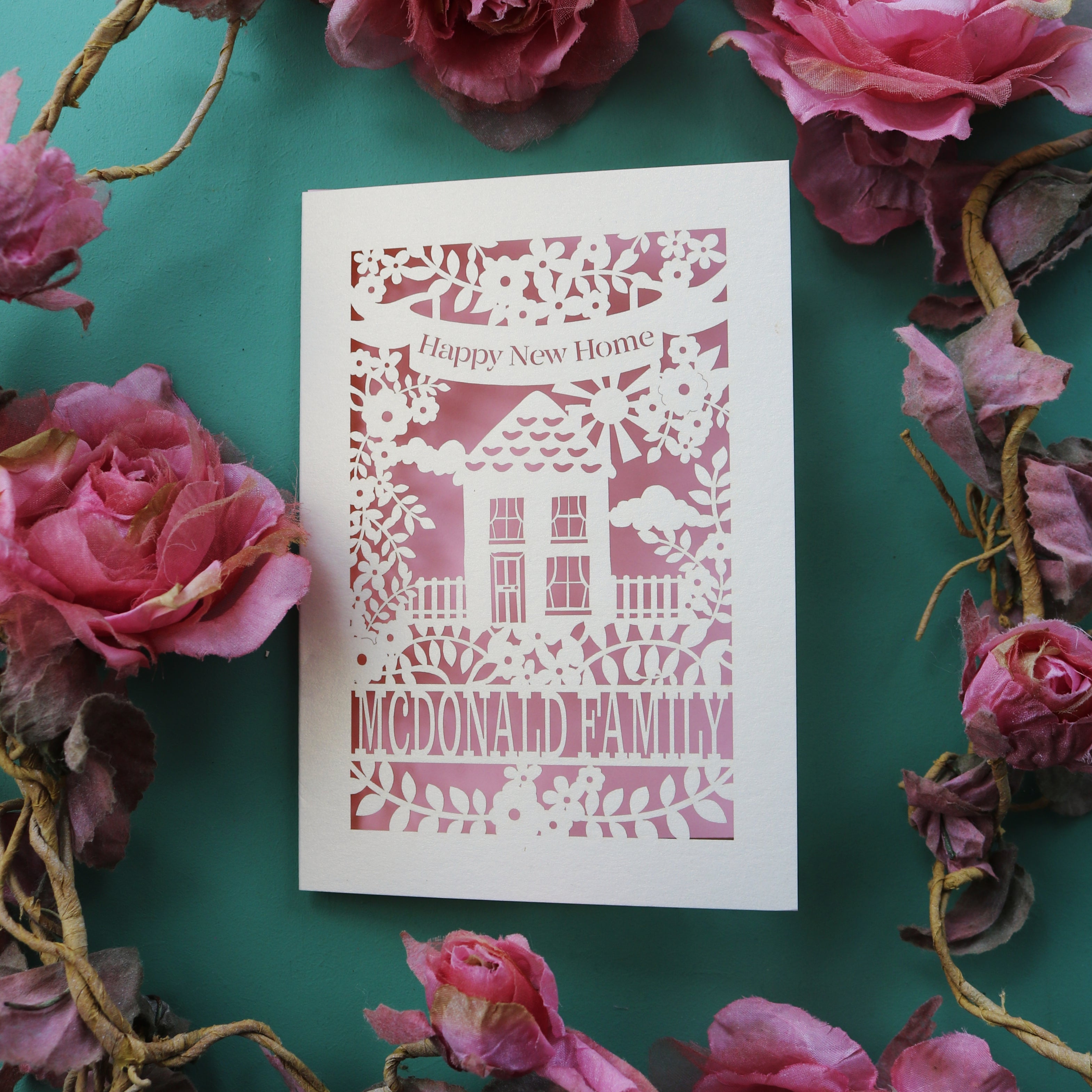 A cream and light pink paper cut house warming card with a house surrounded by flowers. This card reads "happy new home" in a banner 