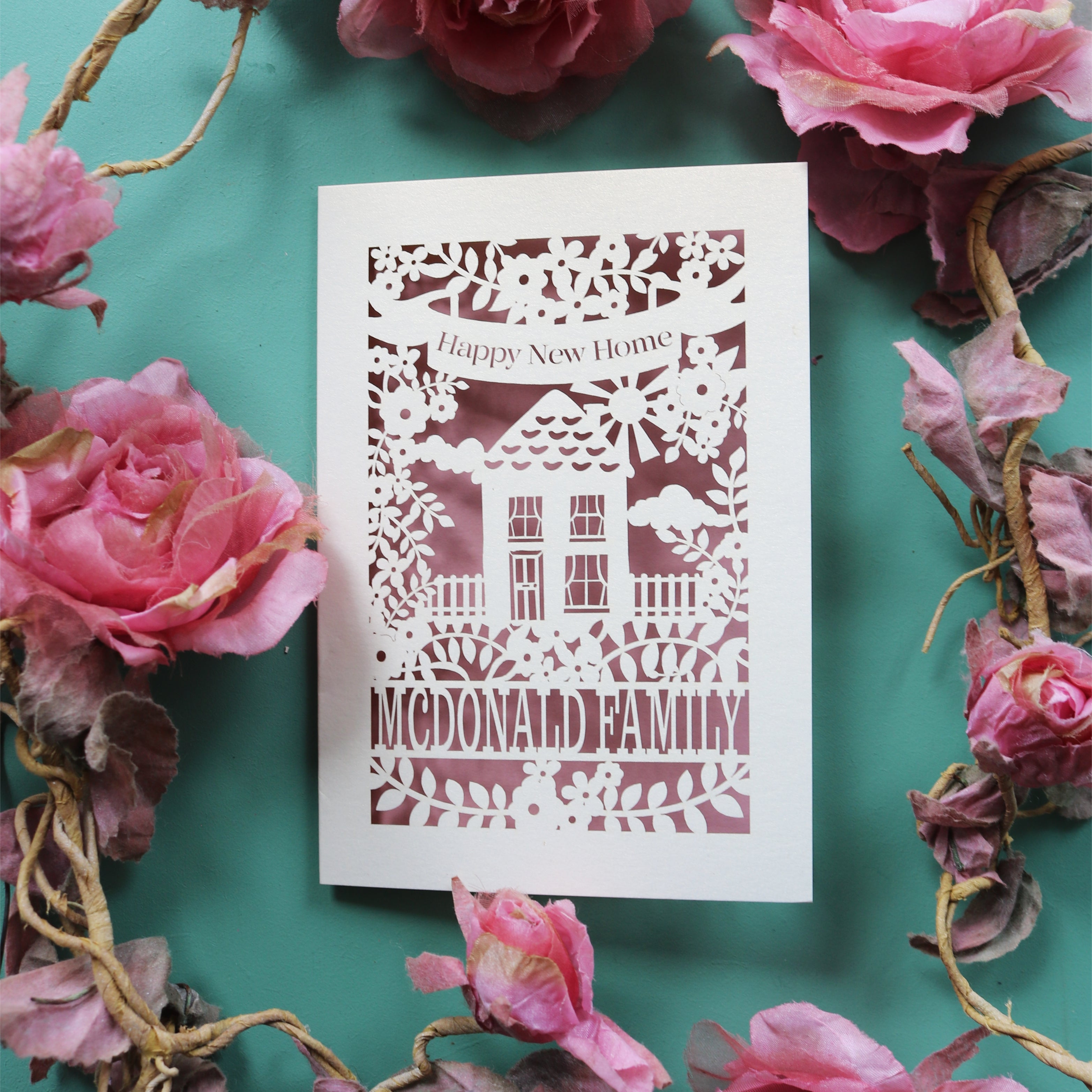 A laser cut card for first time buyers, with a house surrounded by flowers. Card says "Happy new home" and is personalised with the recipient/s' name in a line of text