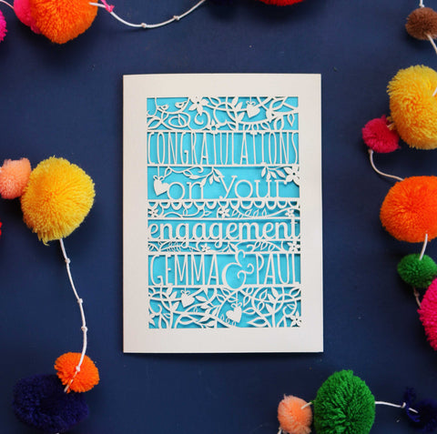 An engagement card personalised and laser cut. Card says "Congratulations on your engagement" and has the couple's names underneath. Card is cut out to reveal a blue paper behind.  - A5 / Cream / Peacock Blue