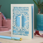 Laser cut 1st Birthday card with cut-out floral design on a desk with stationery items