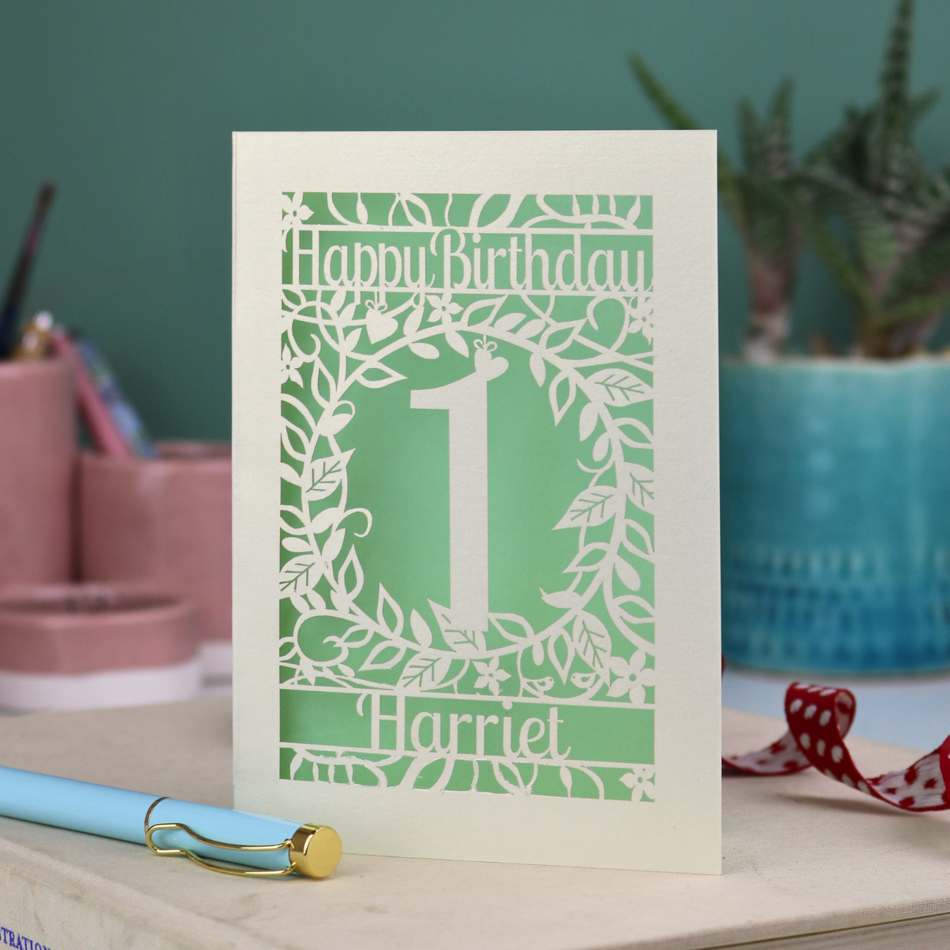 Laser cut personalised 1st Birthday card with cut-out floral design, "Happy Birthday" text, a number 1 and a name.  on a desk with stationery items