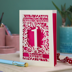 Laser cut 1st Birthday card with cut-out floral design on a desk with stationery items