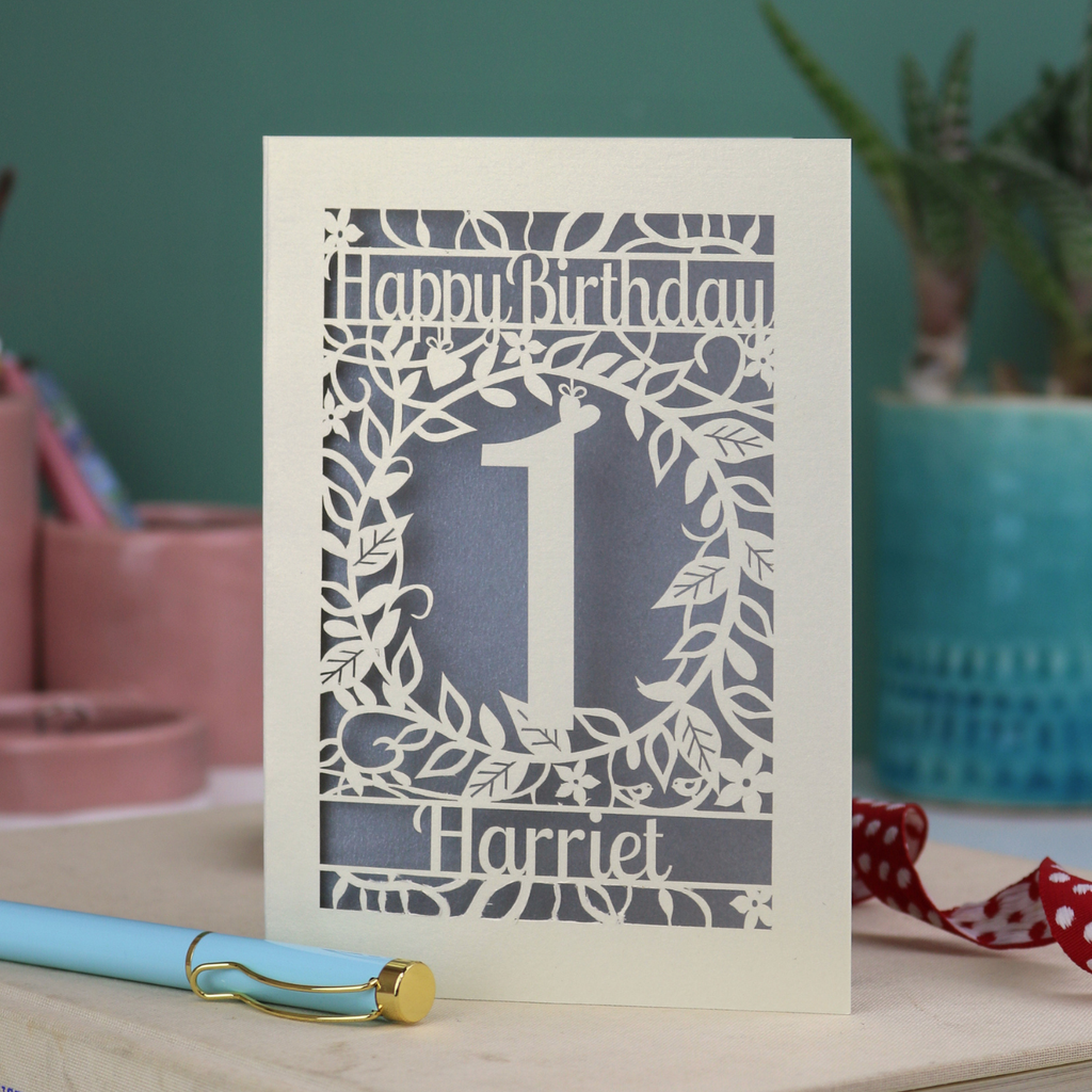 First Birthday card with cut-out design and 'Happy Birthday Harriet' text on a table with plants in the background.