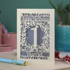 First Birthday card with cut-out design and 'Happy Birthday Harriet' text on a table with plants in the background.