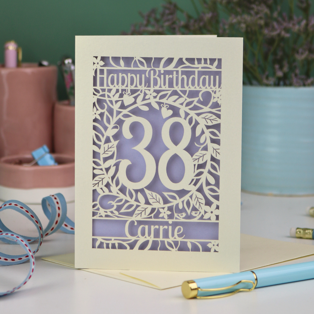A paper cut birthday card, personalised with the age 38 and the name Carrie, with floral border in cream and a coloured paper insert. 