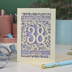 A paper cut birthday card, personalised with the age 38 and the name Carrie, with floral border in cream and a coloured paper insert. 