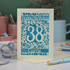 A laser cut birthday card, personalised with the age 38 and the name Carrie, with floral border in cream and a coloured paper insert. 