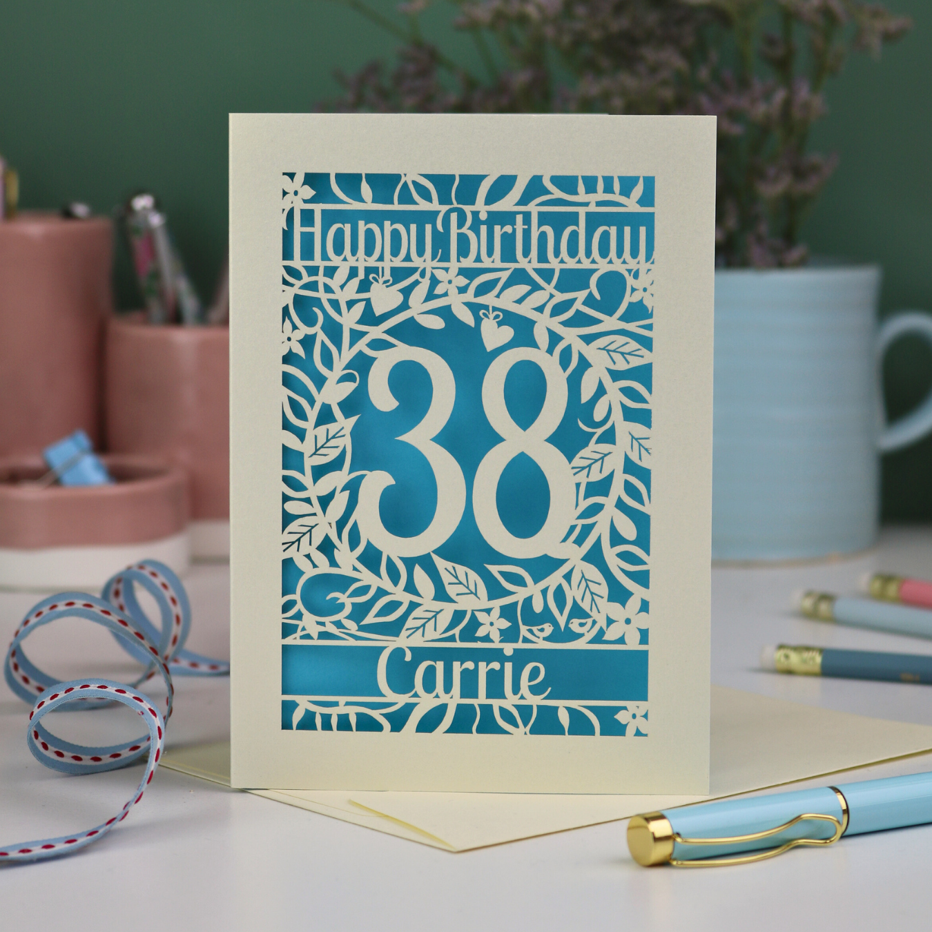 A laser cut birthday card, personalised with the age 38 and the name Carrie, with floral border in cream and a coloured paper insert. 