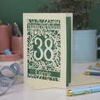 A cut out floral birthday card, personalised with the age 38 and the name Carrie, with floral border in cream and a coloured paper insert. 