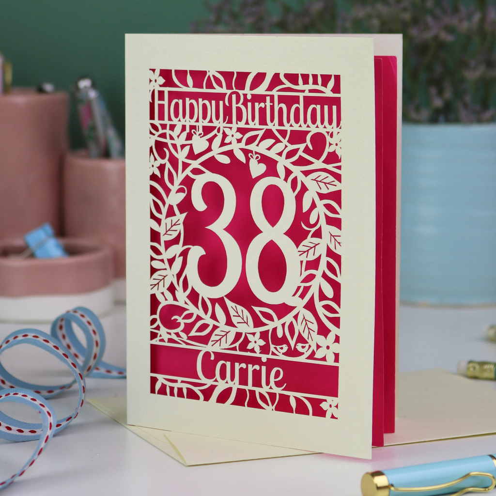 A shocking pink and cream laser cut floral birthday card, personalised with the age 38 and the name Carrie, with floral border in cream and a coloured paper insert. 