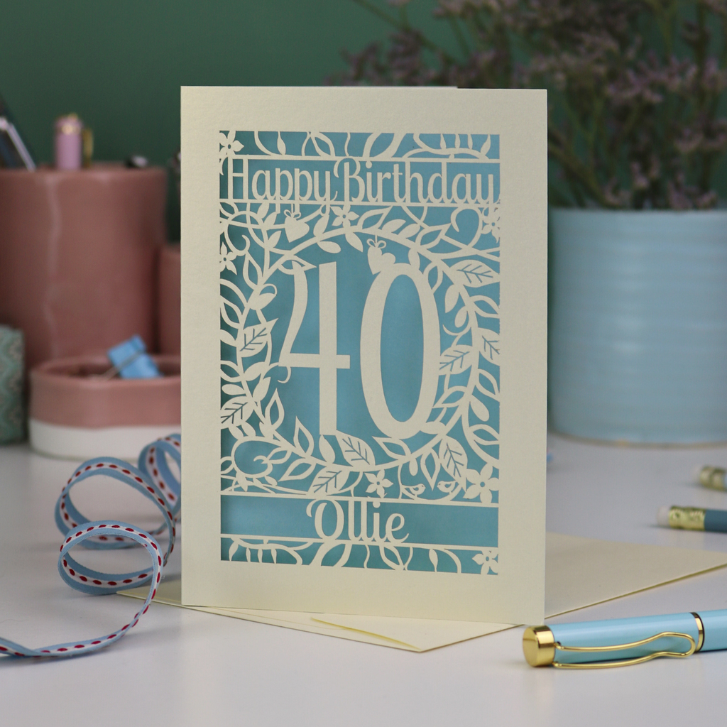 A laser cut floral 40th birthday card, personalised with the number 40 and the name Ollie, on a white desk with stationery items. 