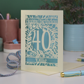 A laser cut floral 40th birthday card, personalised with the number 40 and the name Ollie, on a white desk with stationery items. 