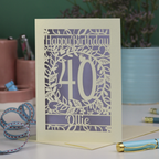 A special laser cut floral 40th birthday card, personalised with the number 40 and the name Ollie, on a white desk with stationery items. 