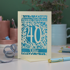 A cut out floral 40th birthday card, personalised with the number 40 and the name Ollie, on a white desk with stationery items. 