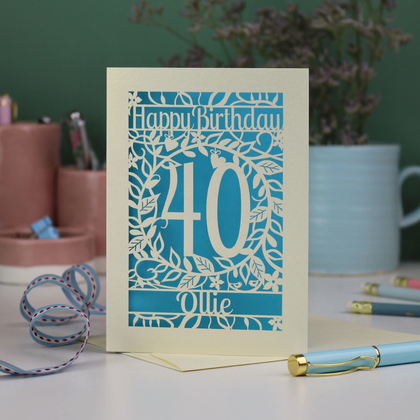 A cut out floral 40th birthday card, personalised with the number 40 and the name Ollie, on a white desk with stationery items. 