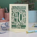 A laser cut personalised 40th birthday card, personalised with the number 40 and the name Ollie, on a white desk with stationery items. 
