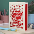 Personalized wedding card with red and cream cut out "forever and always" and floral design, personalised with a date and first names of the couple, on a desk with stationery items.