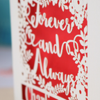 Personalized wedding card with red and cream cut out "forever and always" and floral design, personalised with a date and first names of the couple, on a desk with stationery items.
