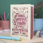 Personalized wedding card with red and cream cut out "forever and always" and floral design, personalised with a date and first names of the couple, on a desk with stationery items.