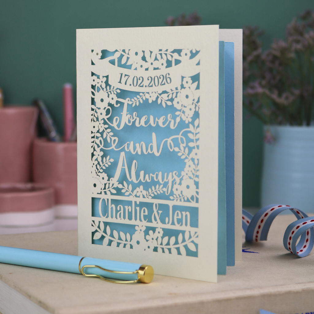 Personalized wedding card with red and cream cut out "forever and always" and floral design, personalised with a date and first names of the couple, on a desk with stationery items.