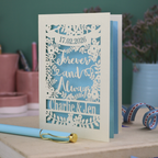 Personalized wedding card with red and cream cut out "forever and always" and floral design, personalised with a date and first names of the couple, on a desk with stationery items.