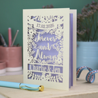 Personalized wedding card with red and cream cut out "forever and always" and floral design, personalised with a date and first names of the couple, on a desk with stationery items.