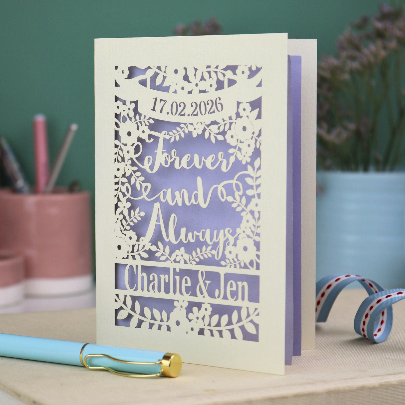 Personalized wedding card with red and cream cut out "forever and always" and floral design, personalised with a date and first names of the couple, on a desk with stationery items.