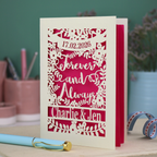 Personalized wedding card with red and cream cut out "forever and always" and floral design, personalised with a date and first names of the couple, on a desk with stationery items.