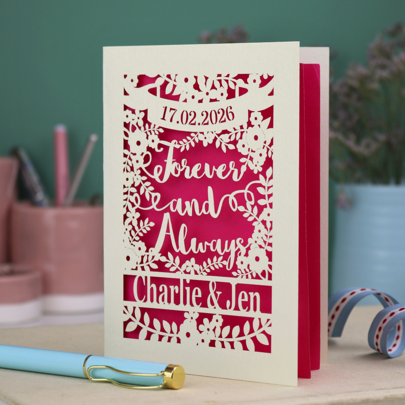 Personalized wedding card with red and cream cut out "forever and always" and floral design, personalised with a date and first names of the couple, on a desk with stationery items.