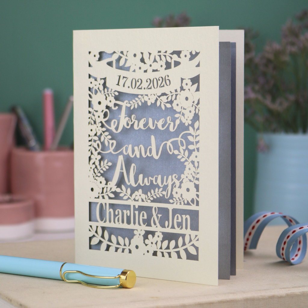 Personalized wedding card with red and cream cut out "forever and always" and floral design, personalised with a date and first names of the couple, on a desk with stationery items.