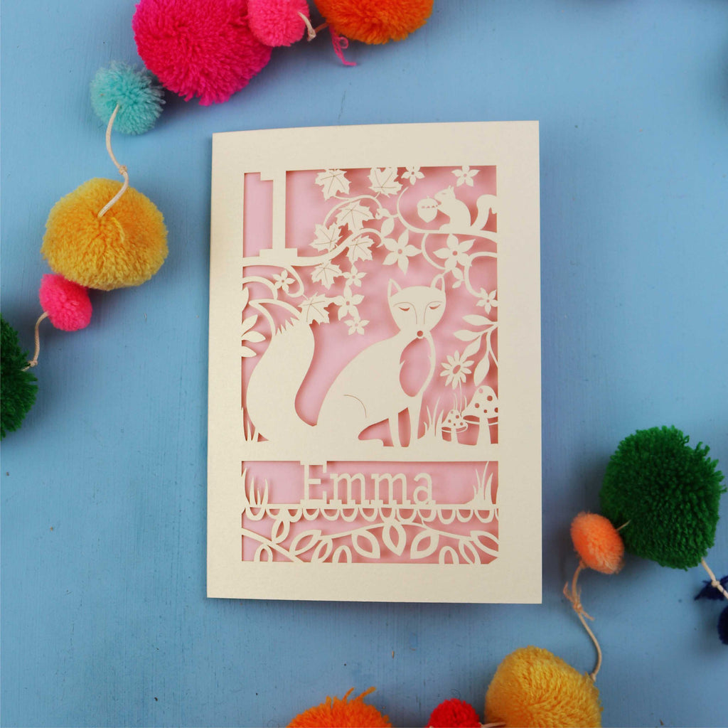 Decorative first birthday card with cut-out design of a fox and floral patterns on a blue background with colourful pom-poms.