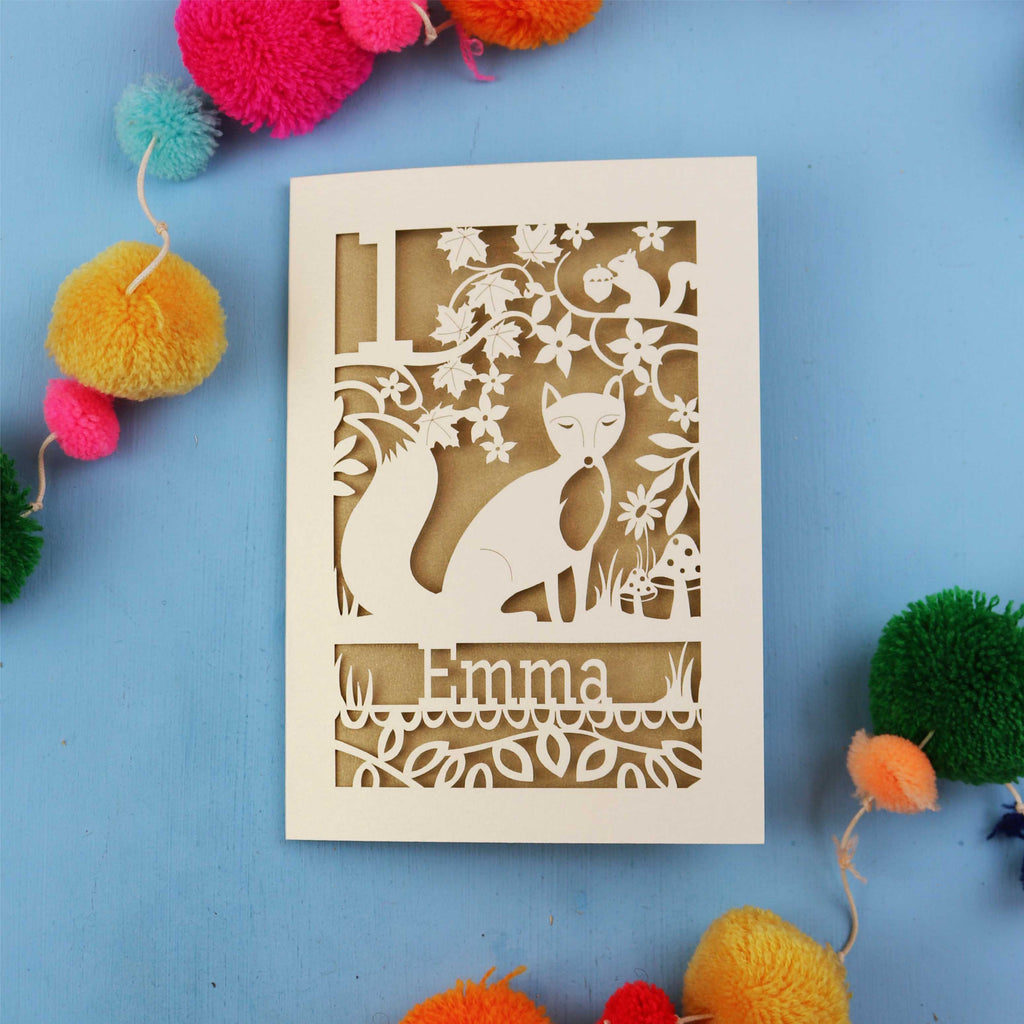 Decorative first birthday card with cut-out design of a fox and floral patterns on a blue background with colourful pom-poms.
