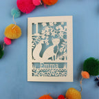Decorative first birthday card with cut-out design of a fox and floral patterns on a blue background with colourful pom-poms.