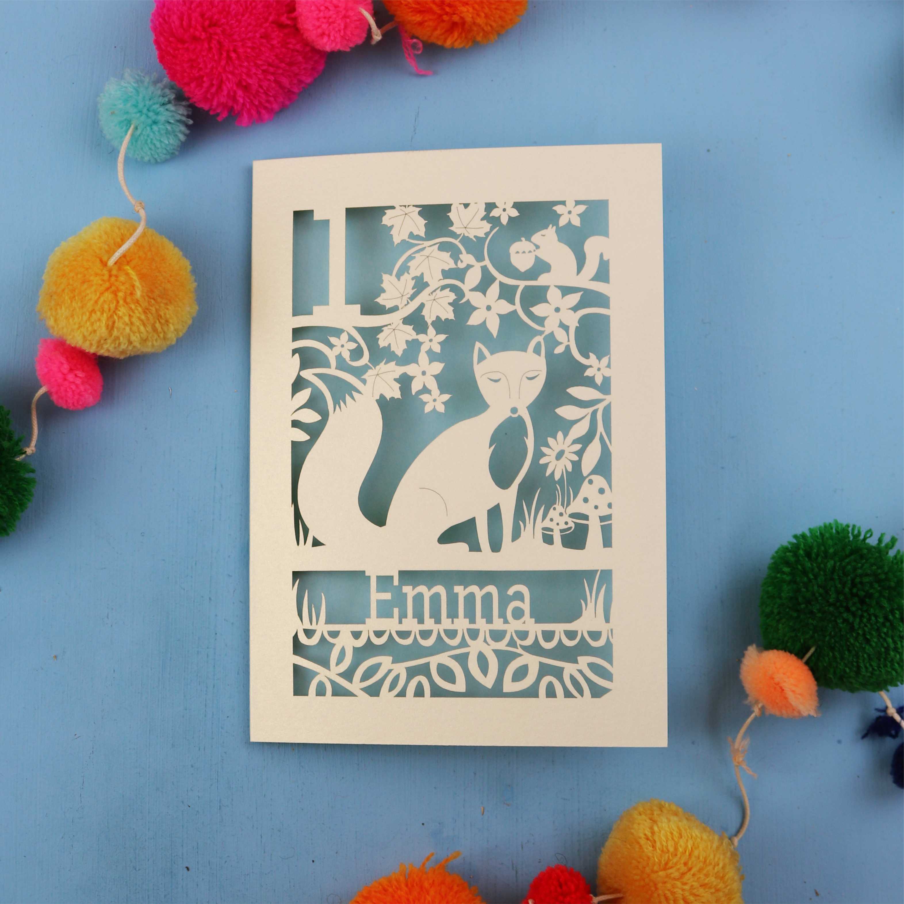 Decorative first birthday card with cut-out design of a fox and floral patterns on a blue background with colourful pom-poms.