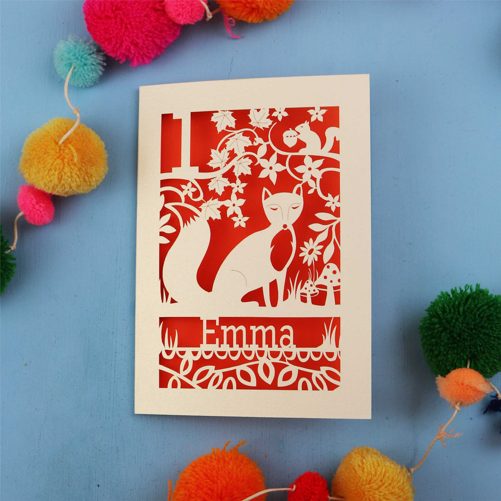 Decorative first birthday card with cut-out design of a fox and floral patterns on a blue background with colourful pom-poms.