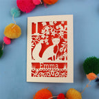 Decorative first birthday card with cut-out design of a fox and floral patterns on a blue background with colourful pom-poms.
