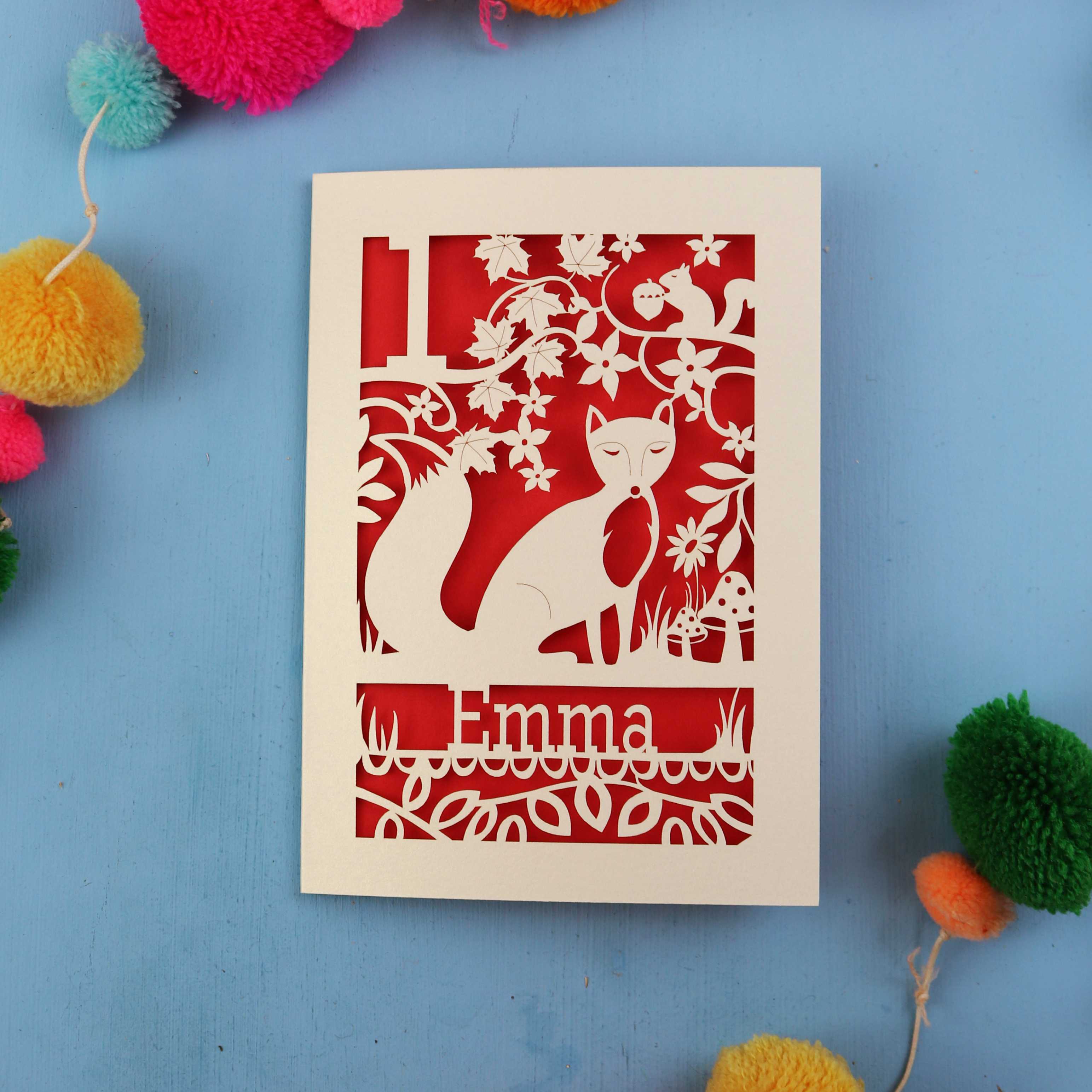 Decorative first birthday card with cut-out design of a fox and floral patterns on a blue background with colourful pom-poms.