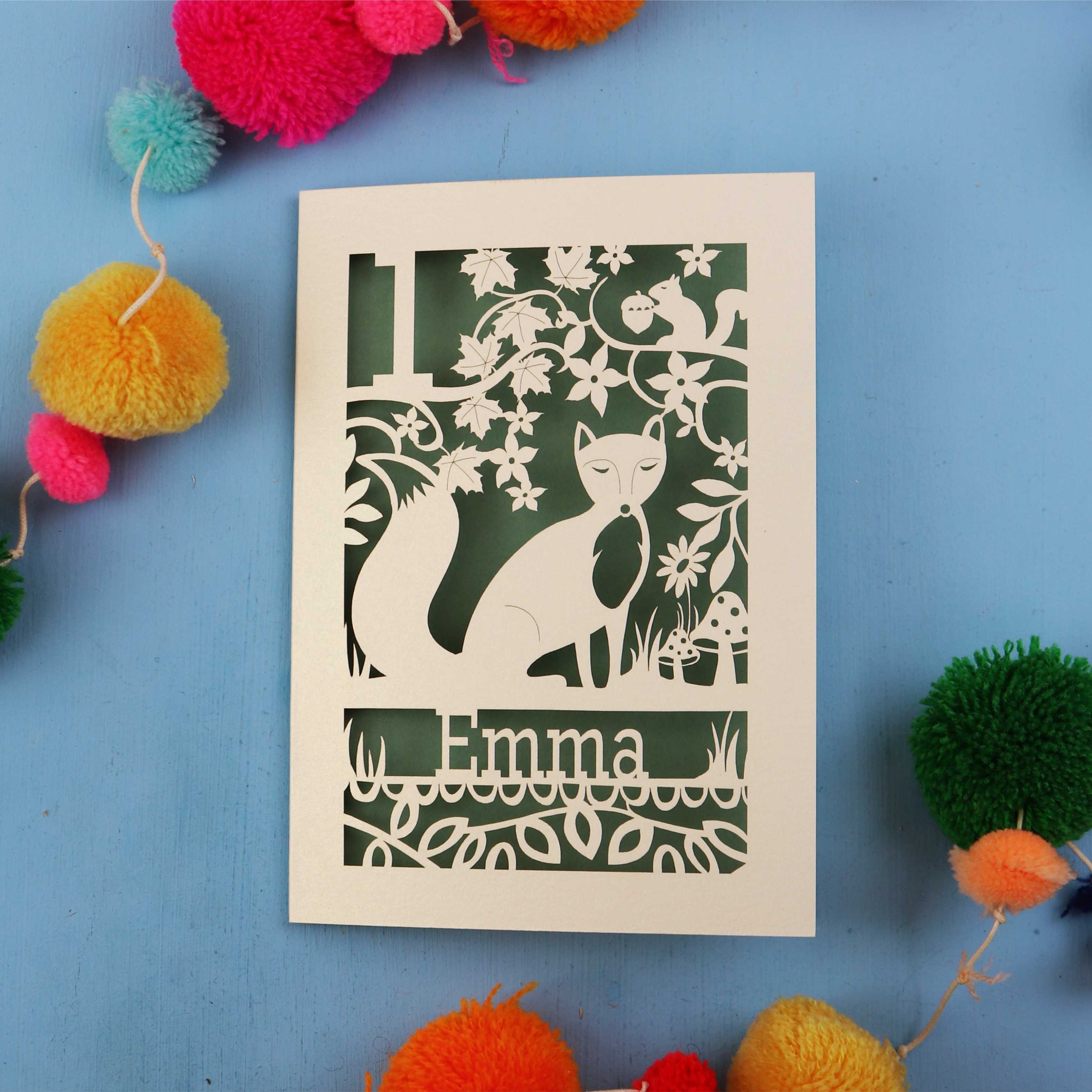 Personalised first birthday card with cut-out design of a fox and floral patterns on a blue background with colourful pom-poms.