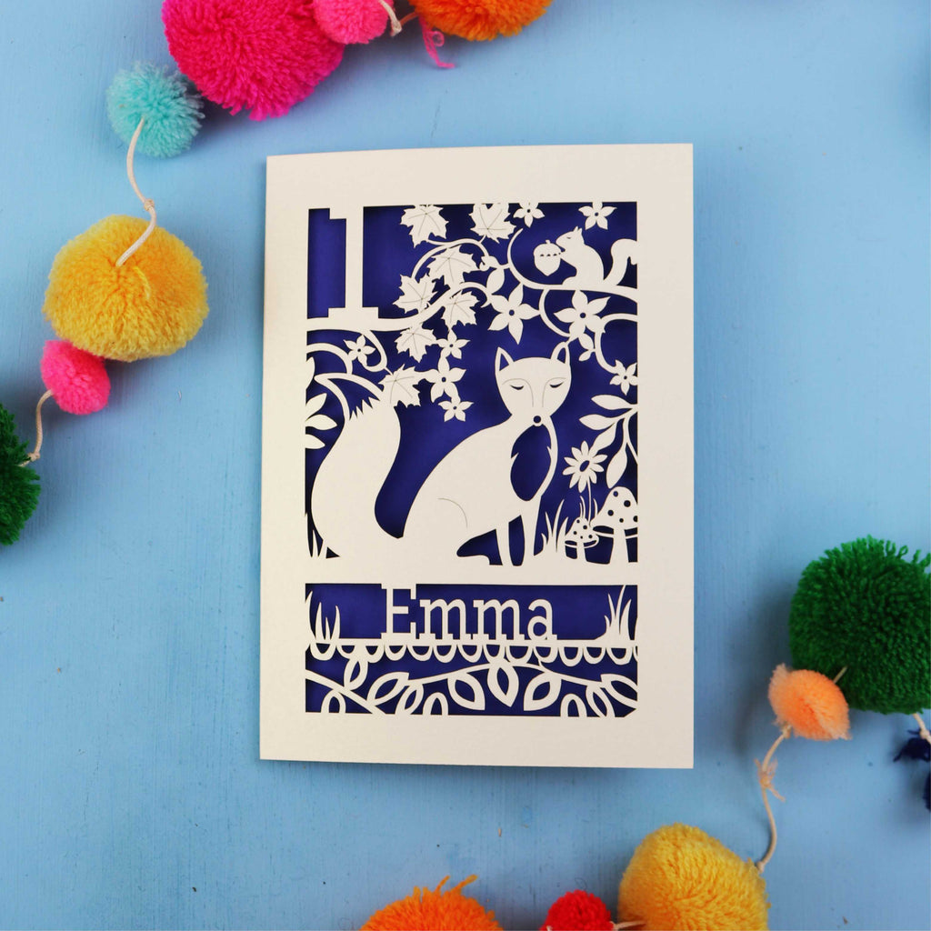 Laser cut Personalised first birthday card with cut-out design of a fox and floral patterns on a blue background with colourful pom-poms.