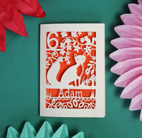 A laser cut birthday card, personalised with a name and age. There is a fox in the centre of the design, with leaf and flower shapes around. This one has a bright orange paper insert.  - 