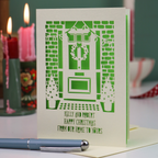 A handmade Christmas card with laser-cut illustration of a front door, wreath, brick wall, and small trees, with the text 'KELLY AND ROBERT HAPPY CHRISTMAS FROM OUR HOME TO YOURS'. The card is standing on a table with a blue pen in the foreground and blurred red candles in the background.
