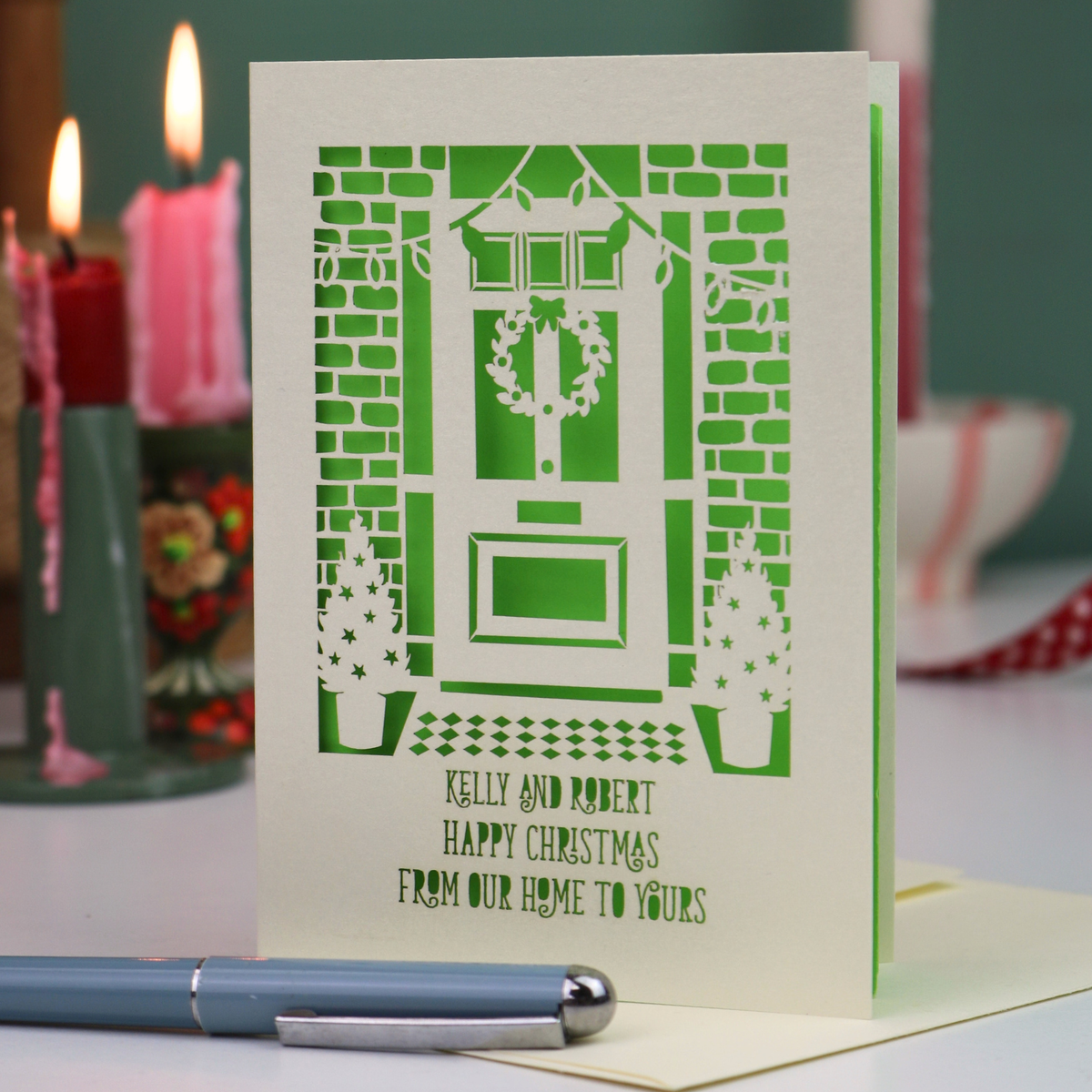 A handmade Christmas card with laser-cut illustration of a front door, wreath, brick wall, and small trees, with the text 'KELLY AND ROBERT HAPPY CHRISTMAS FROM OUR HOME TO YOURS'. The card is standing on a table with a blue pen in the foreground and blurred red candles in the background.