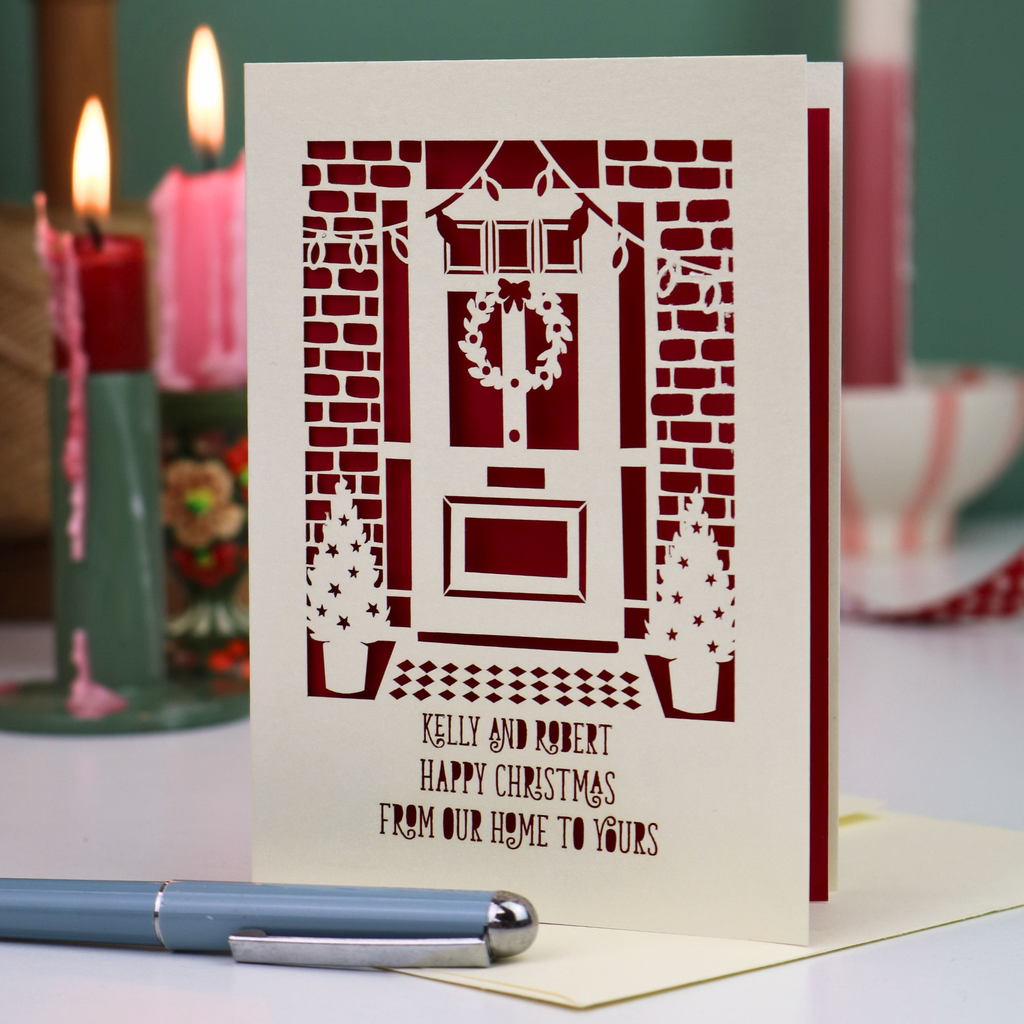 A personalised Christmas card with a laser-cut design of a front door, wreath, and small Christmas trees, displayed on a table with a pen in the foreground and lit candles in the background.